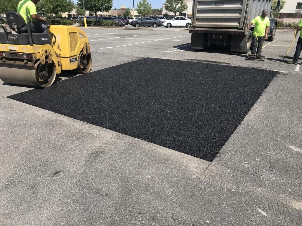 Saw-cut asphalt section removed and replaced with fresh asphalt, compacted by a roller in a commercial parking lot